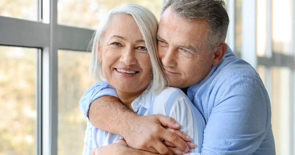 Partnersuche: ein Mann umarmt lachend eine Frau am Fenster