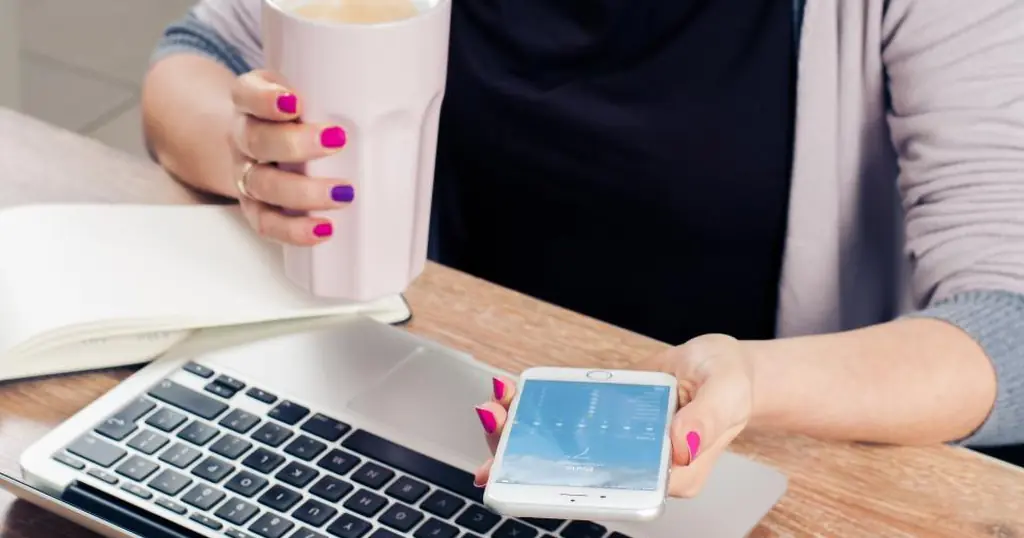 Singlebörse: eine Frau hält am Laptop ein Smartphone und einen Becher in der Hand